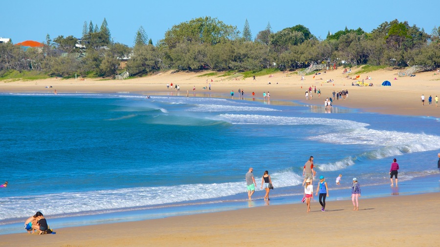 A beach with clear blue water, people walking along the shore, and trees in the background.