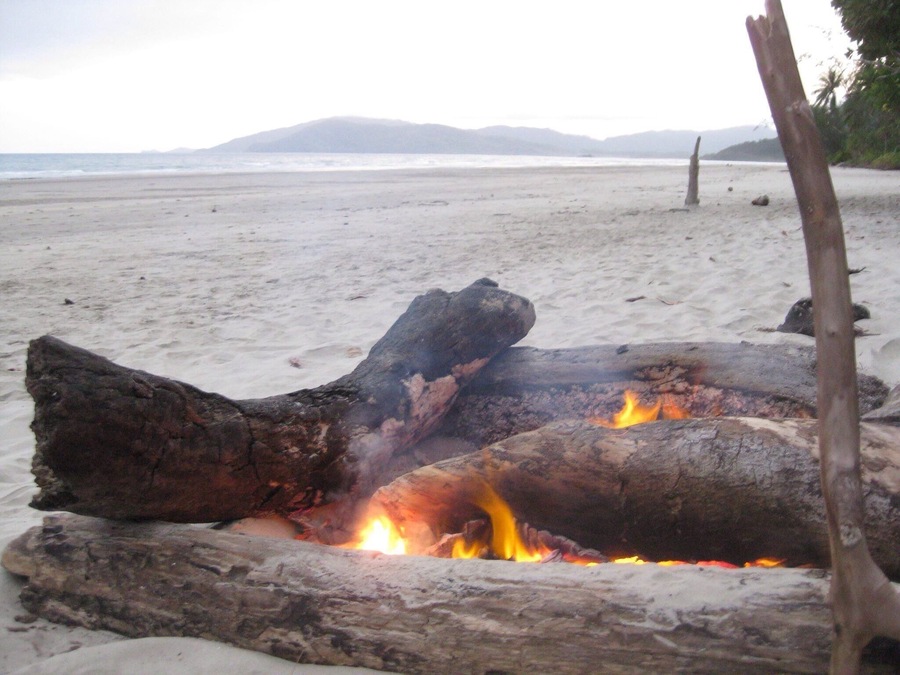 Ein Lagerfeuer an einem sandigen Strand, im Hintergrund ein Berg.