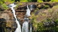 A multi-tiered waterfall cascading over rocky terrain surrounded by lush greenery.