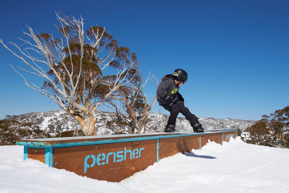 A snowboarder performing a trick on a wooden rail with 'perisher' written on it, surrounded by snow-covered trees and hills.