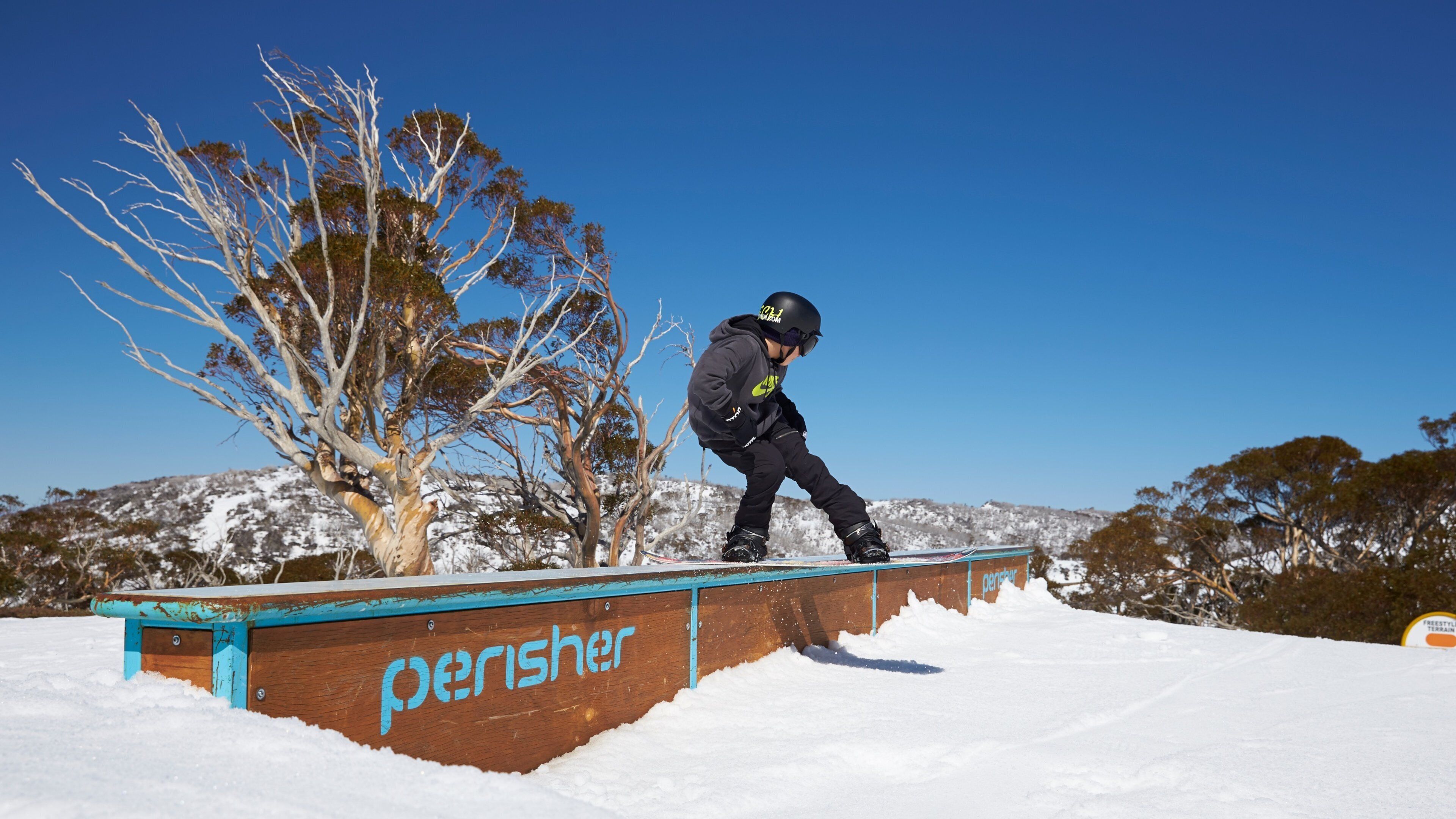 A snowboarder performing a trick on a wooden rail with 'perisher' written on it, surrounded by snow-covered trees and hills.