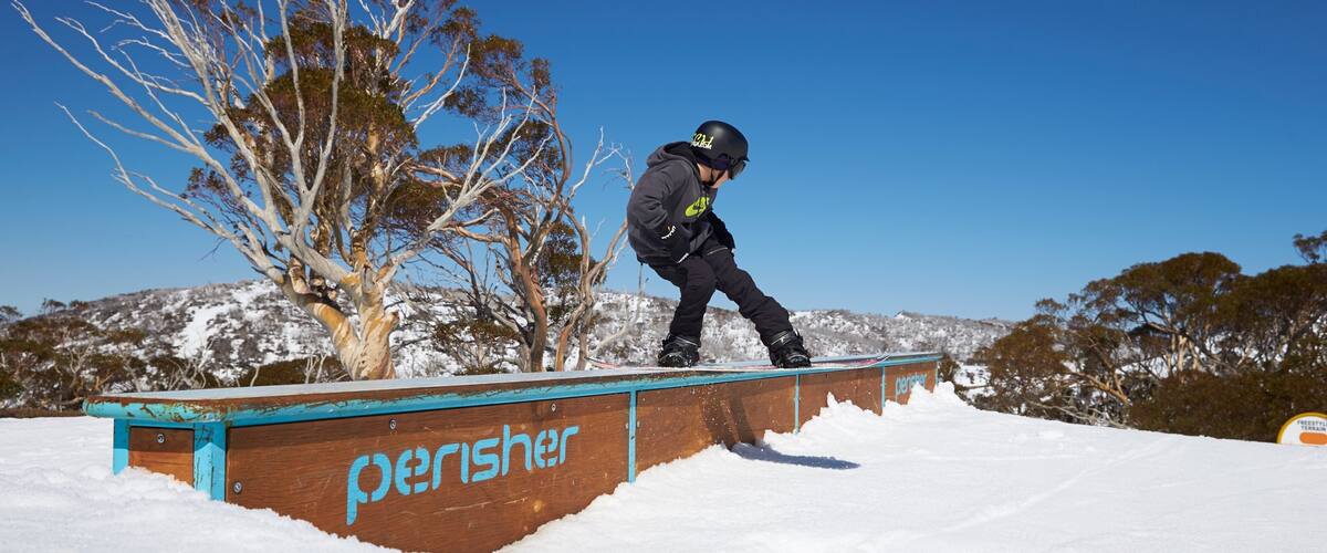 A snowboarder performing a trick on a wooden rail with 'perisher' written on it, surrounded by snow-covered trees and hills.