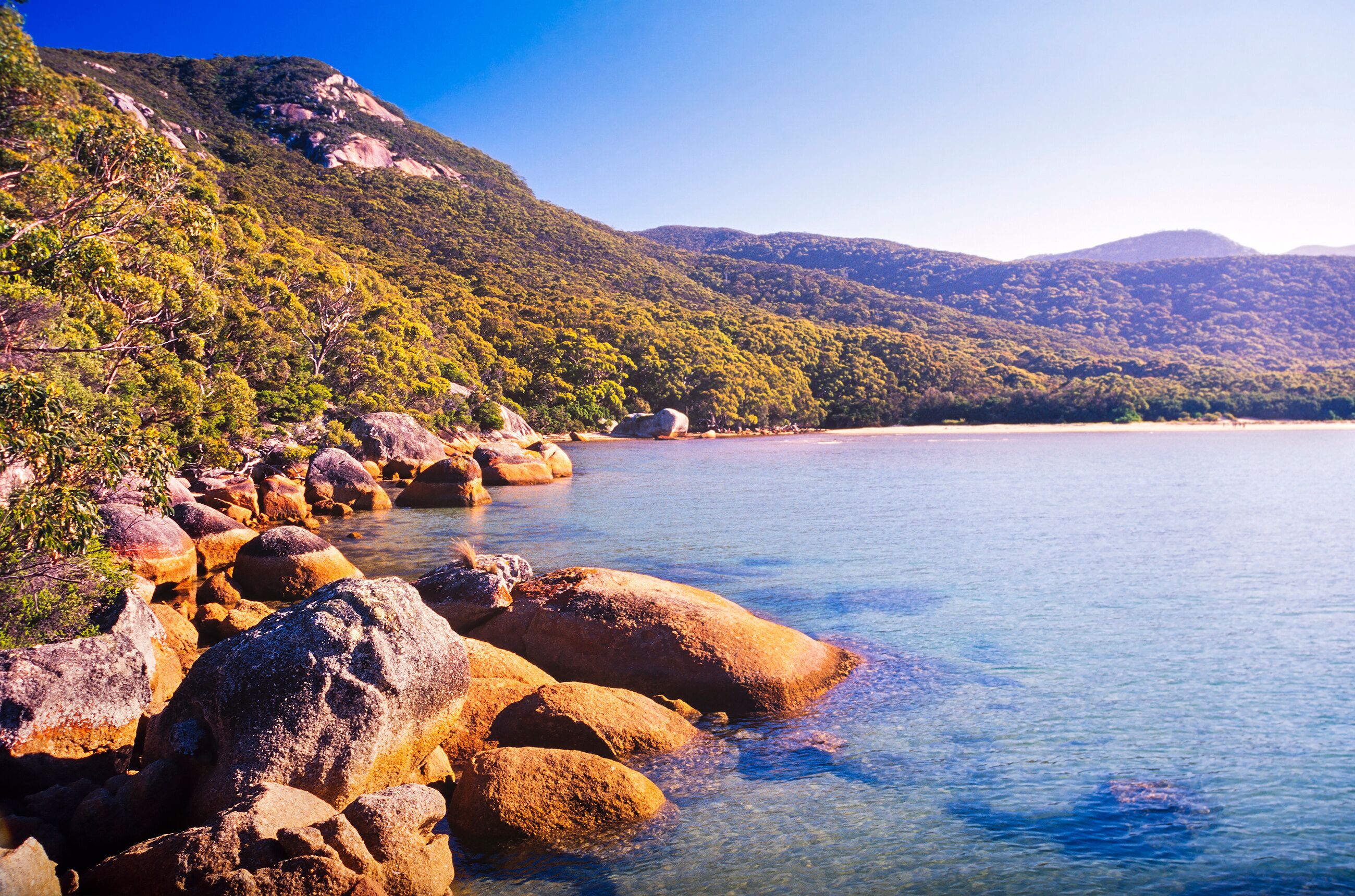 A coastal landscape with large rocks, clear blue water, and green hills.