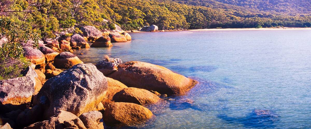 A coastal landscape with large rocks, clear blue water, and green hills.