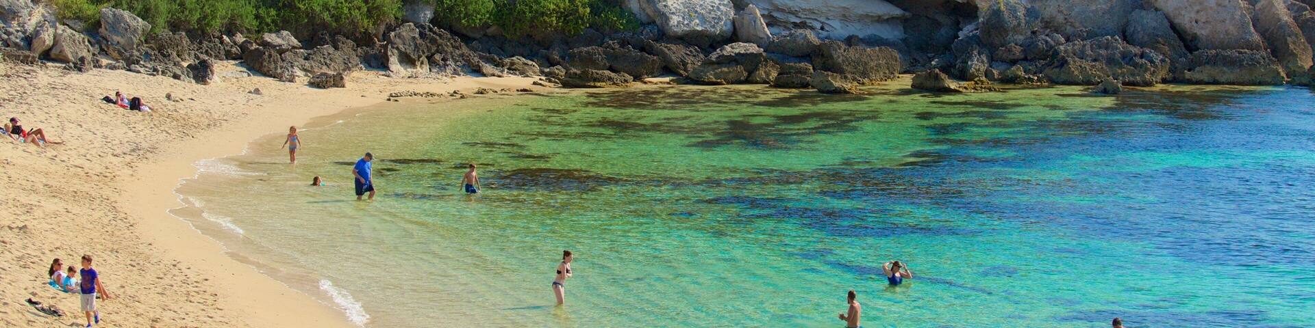 A beach with clear turquoise water, sandy shore, and rocky cliffs with greenery.
