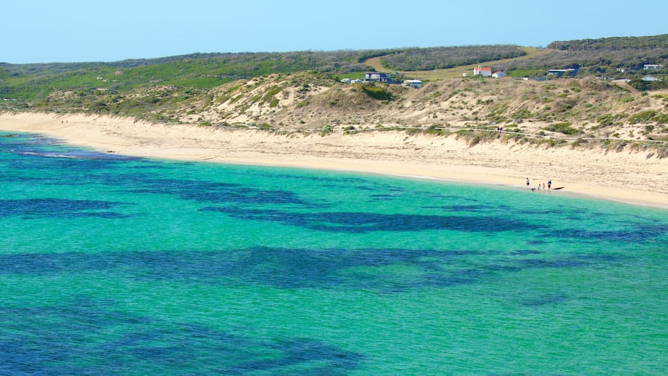 A coastal landscape with clear turquoise waters, sandy beach, and a hilly background with scattered houses.