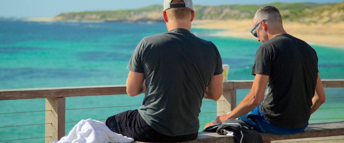 Two men sitting on a wooden bench overlooking a beach with clear turquoise waters and a distant shoreline.