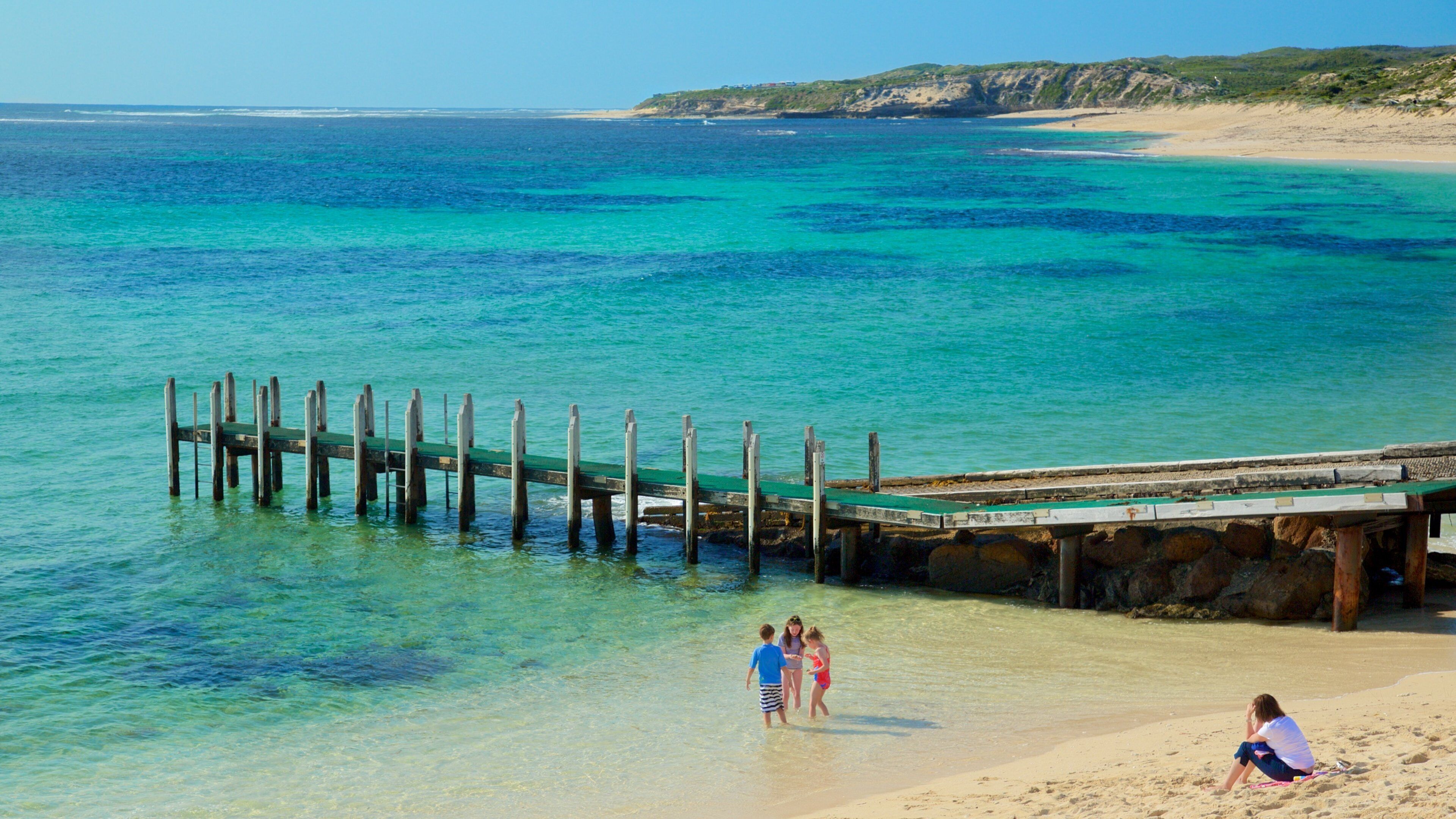 A pier extending into turquoise waters with a beach and rocky cliffs in the background.