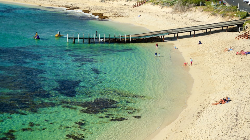 A beach with clear turquoise water, a wooden pier, and people relaxing on the sand.