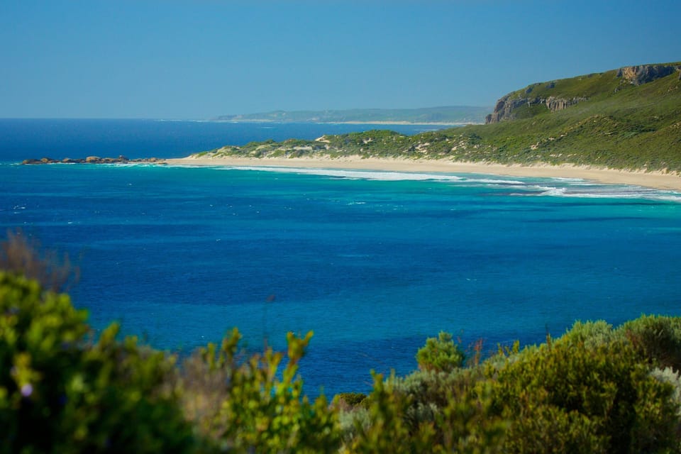 A coastal landscape with a clear blue sea, sandy beach, and green hills.