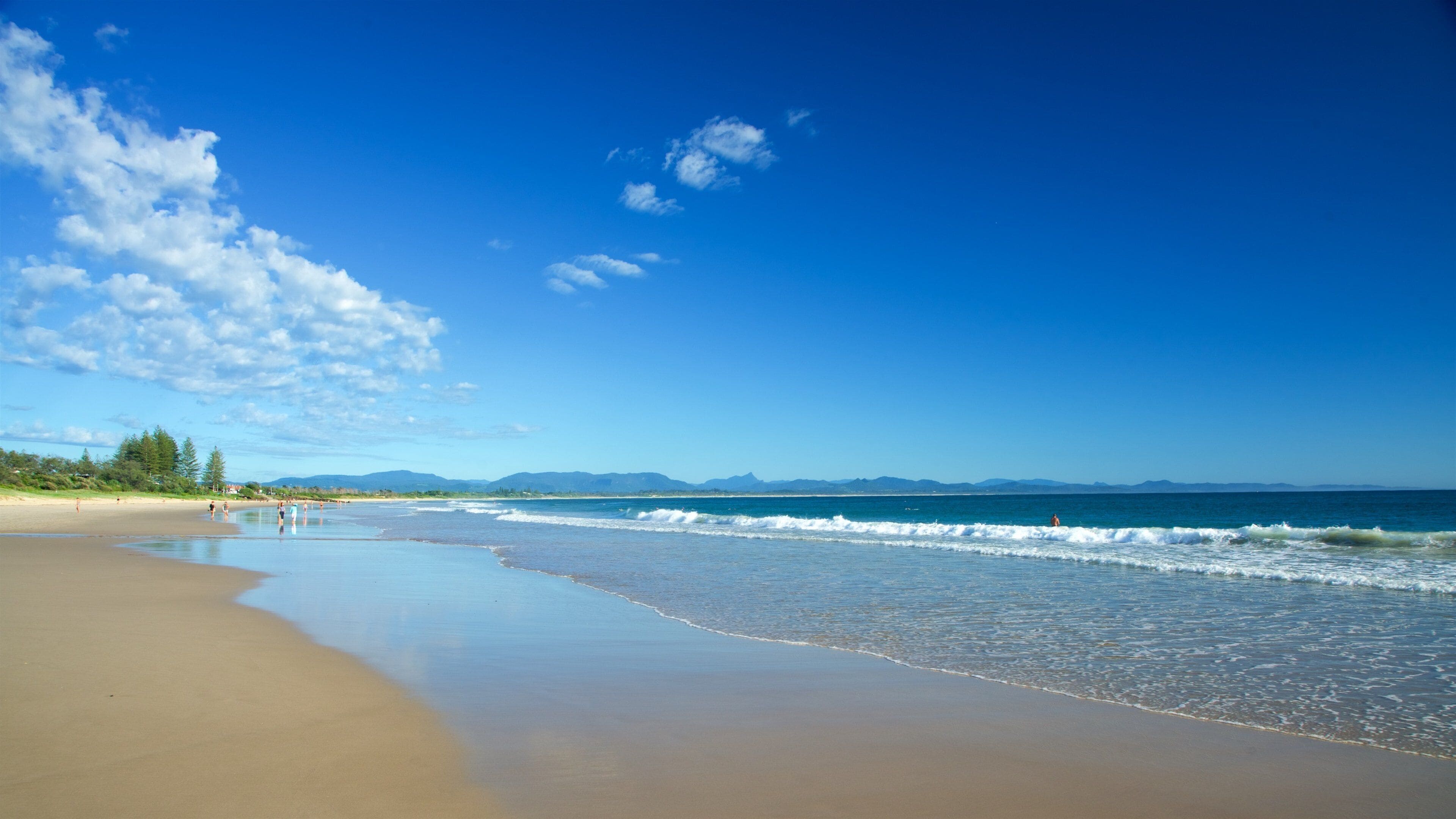 Une plage à l’eau d’un bleu limpide, avec un rivage sablonneux et, au loin, des montagnes sous un ciel partiellement nuageux.