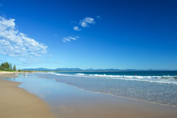 A beach with clear blue water, a sandy shore, and distant mountains under a partly cloudy sky.