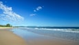 A beach with clear blue water, a sandy shore, and distant mountains under a partly cloudy sky.