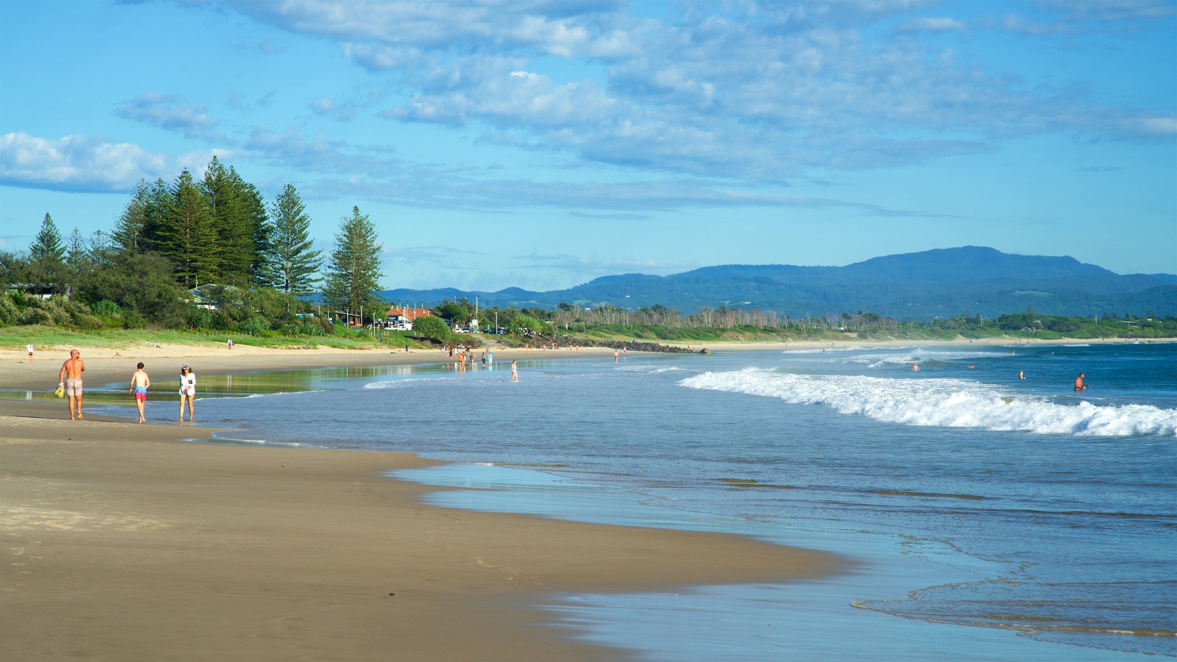 A beach with people walking along the shore, waves crashing onto the sand, and a forested area in the background.