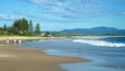 A beach with people walking along the shore, waves crashing onto the sand, and a forested area in the background.