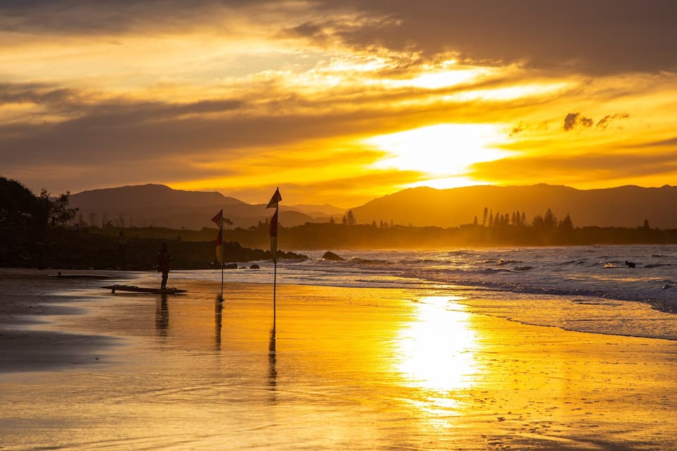 A beach at sunset with waves, a flag, and mountains in the background.