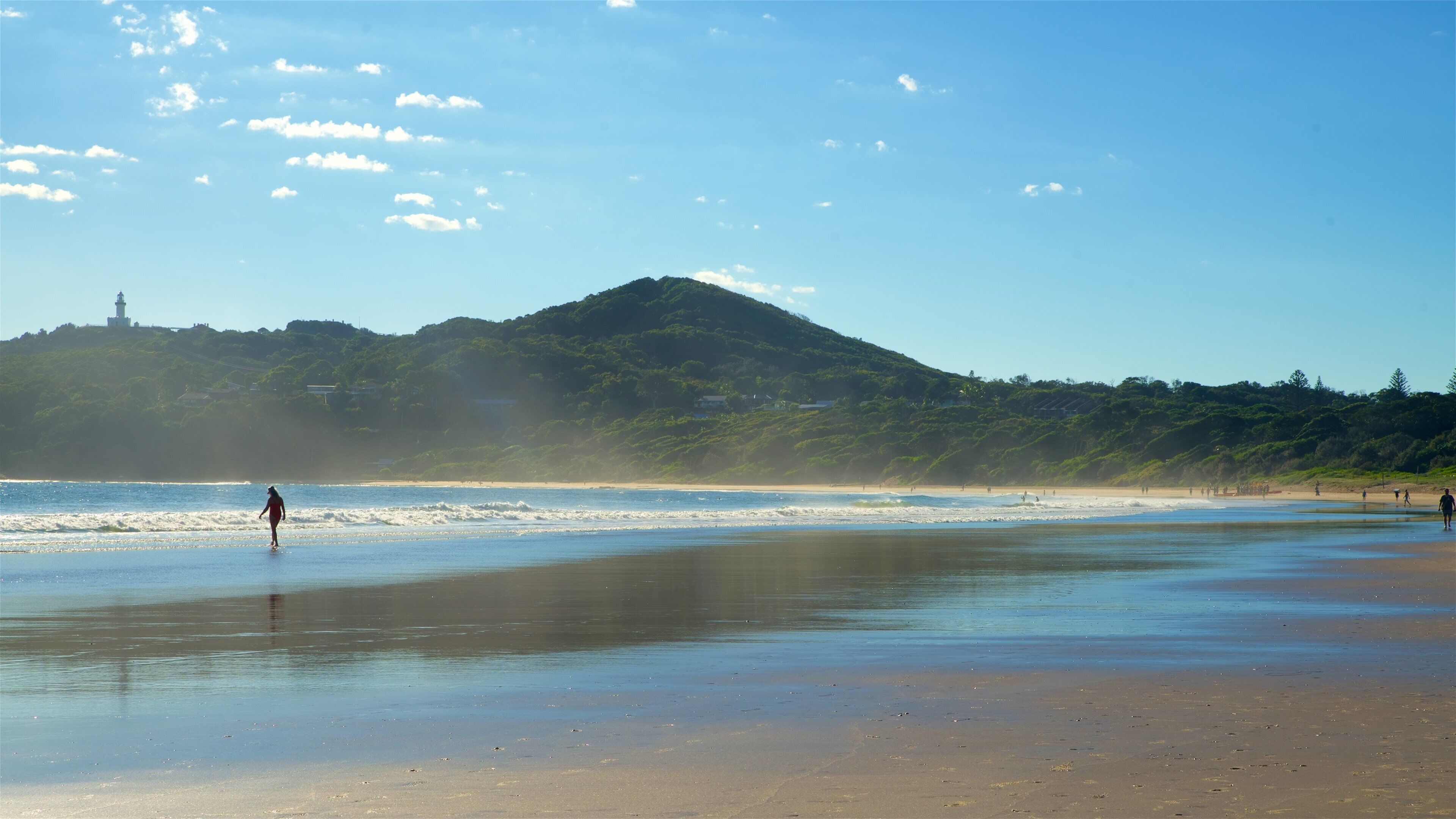 A beach with waves, a lighthouse, and a hill with trees.