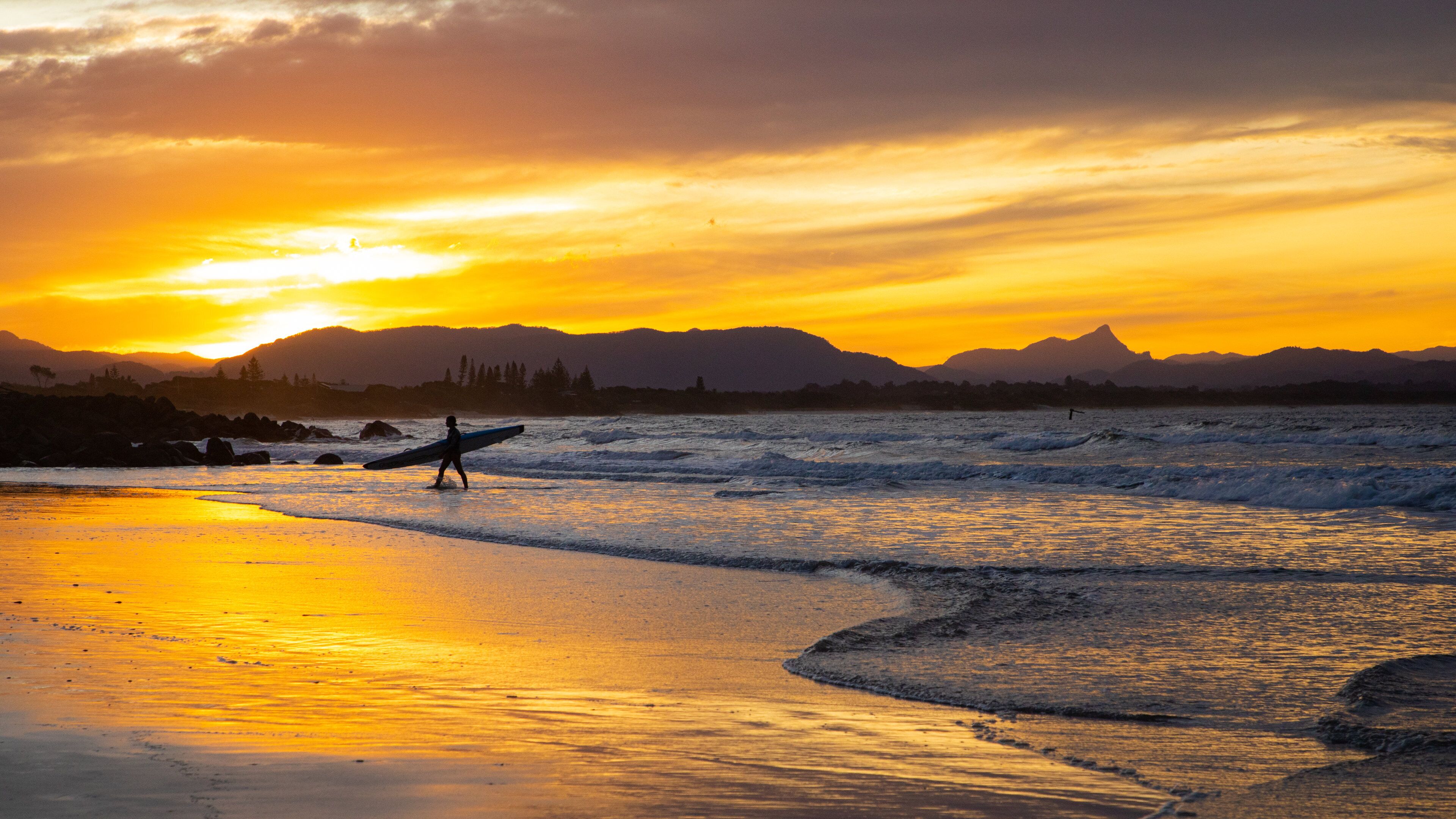 A sunset over a beach with waves, mountains in the background, and a person with a surfboard.