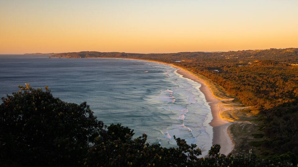 A coastal landscape with a beach, ocean, and forested hills during sunset.