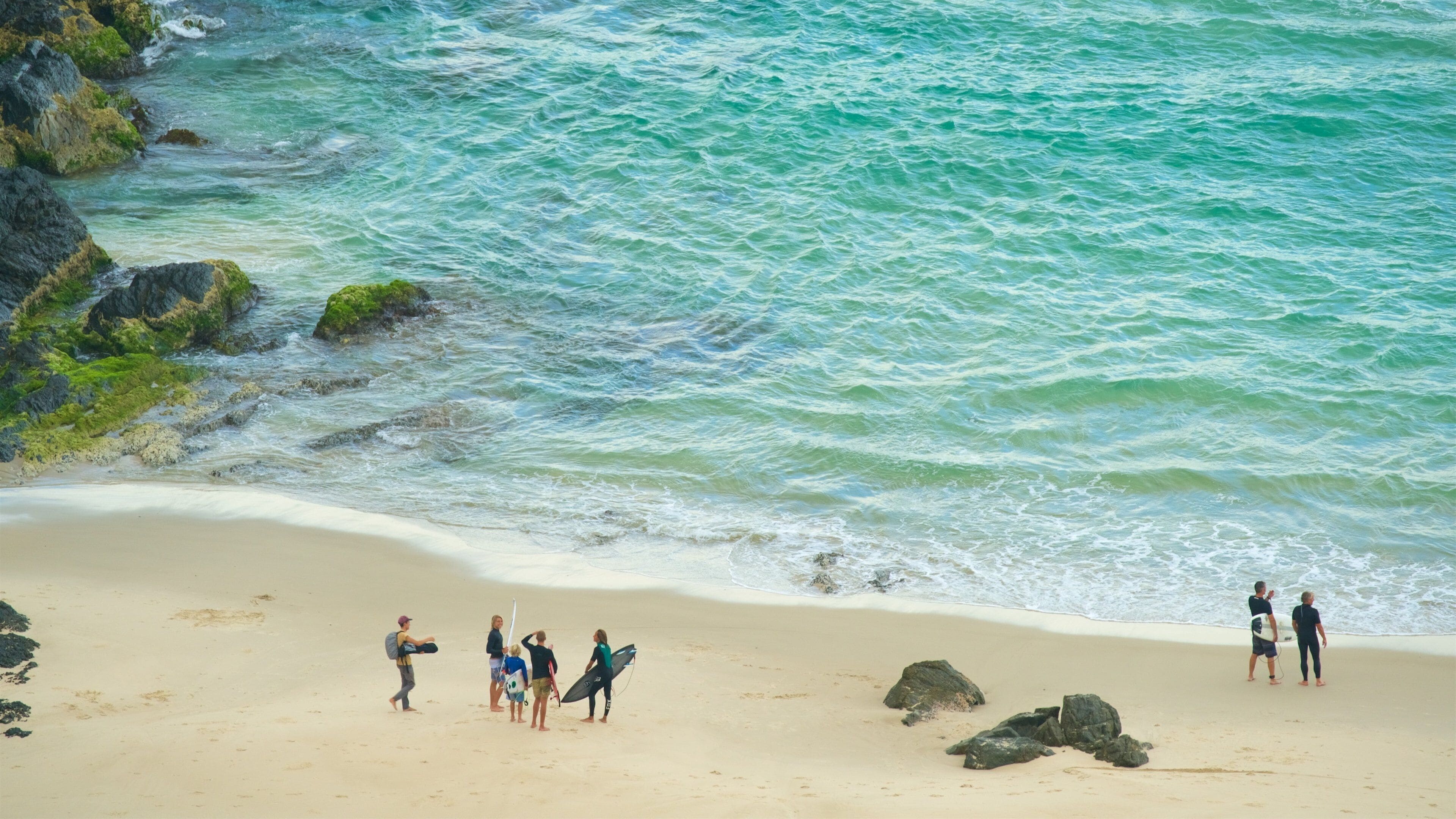 A beach with turquoise water, rocky outcrops, and people walking along the shore.