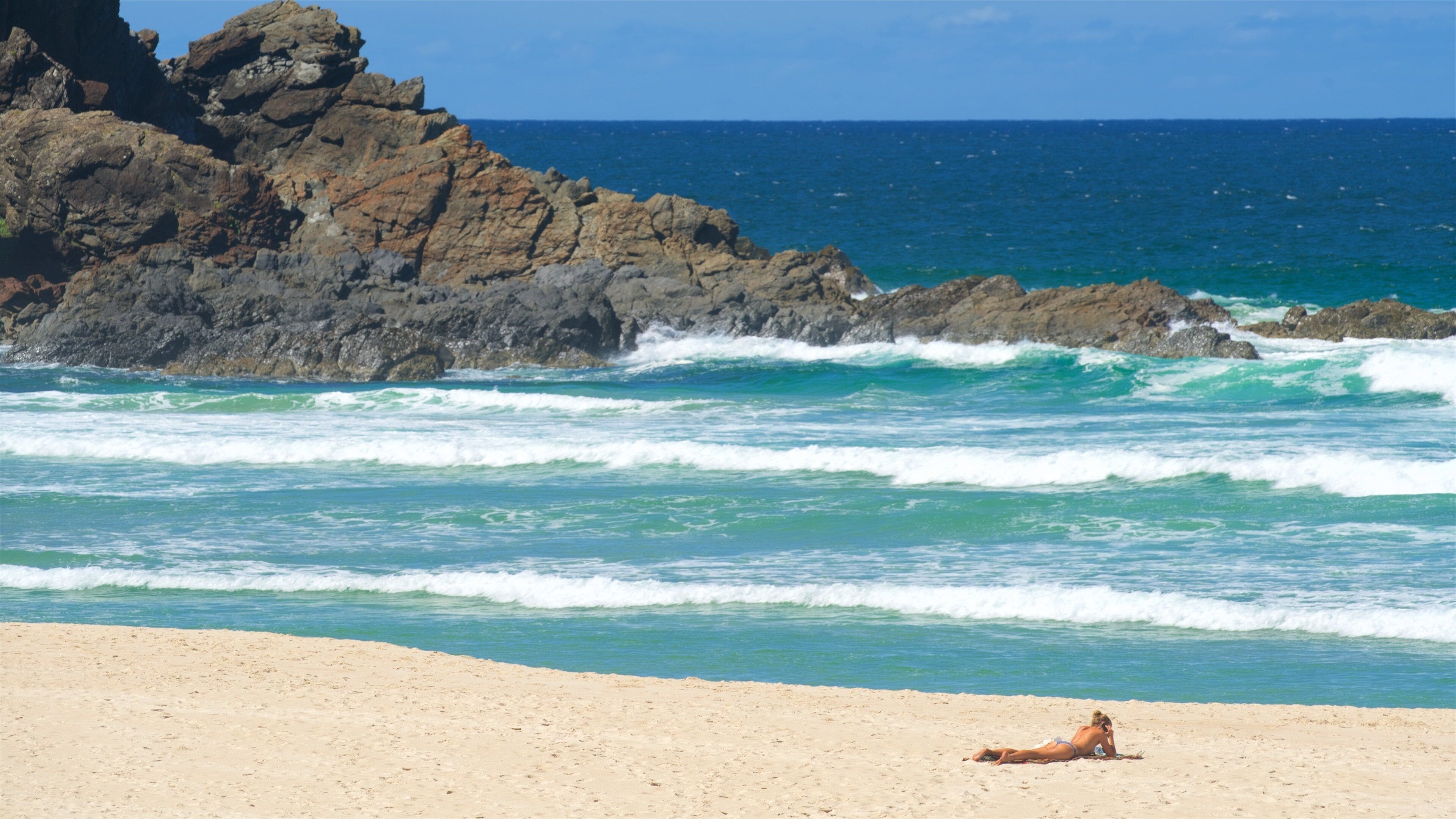 A beach with waves crashing against rocky cliffs and a clear blue sky.
