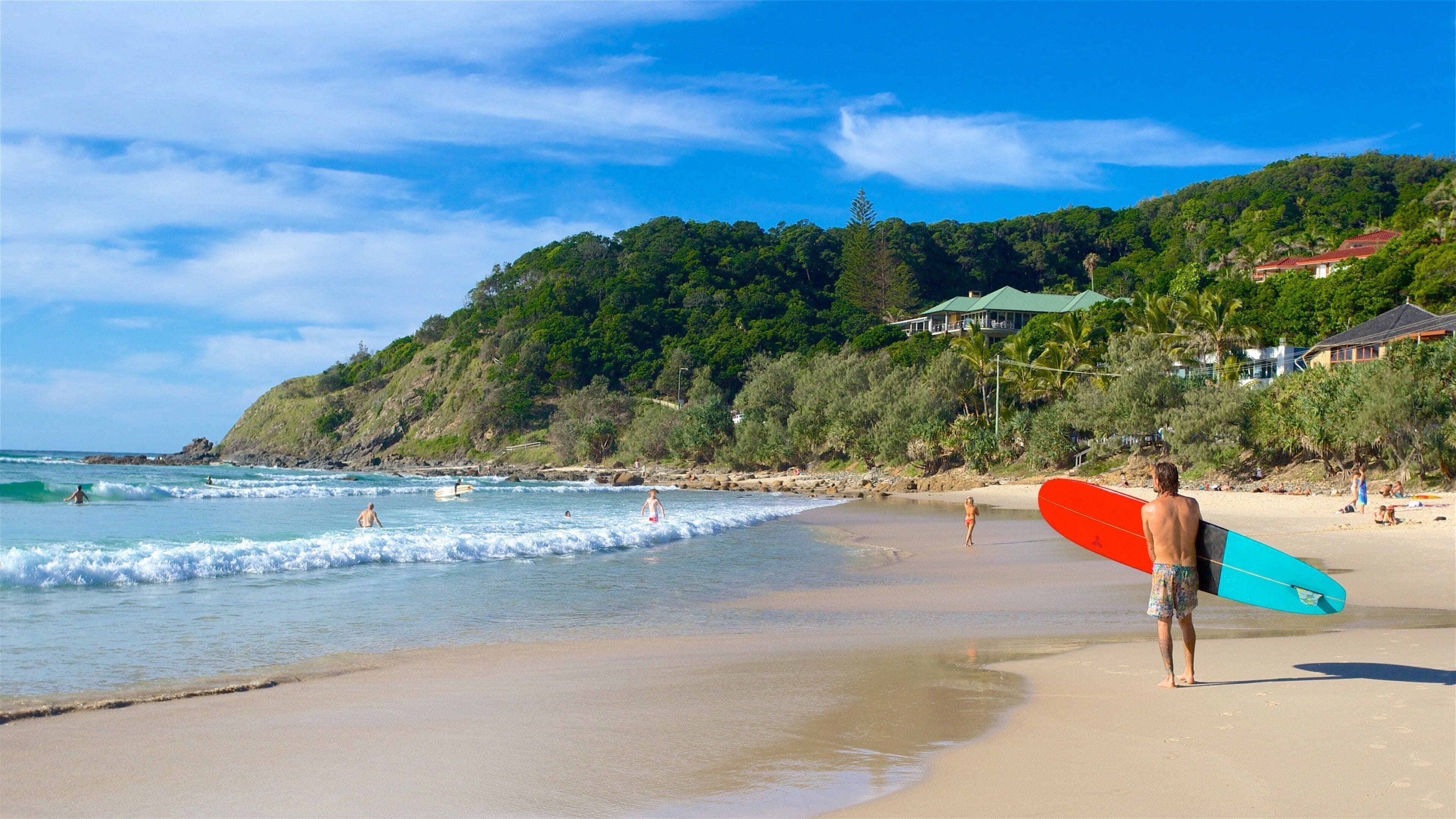 A beach with waves, a person holding a surfboard, and houses on a hillside.