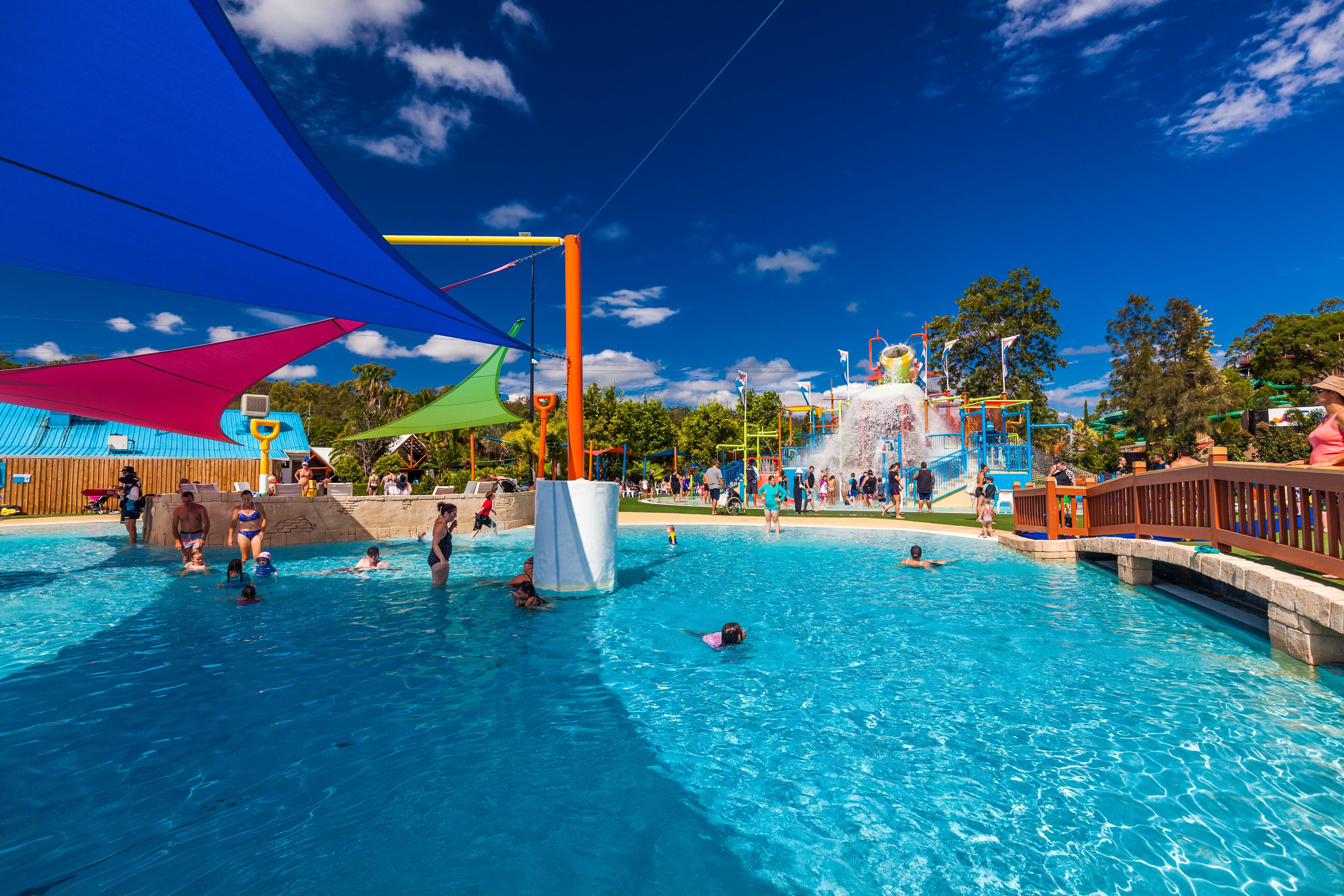 Un parc aquatique animé, doté d’une grande piscine, de jeux colorés et d’un pont enjambant l’eau.