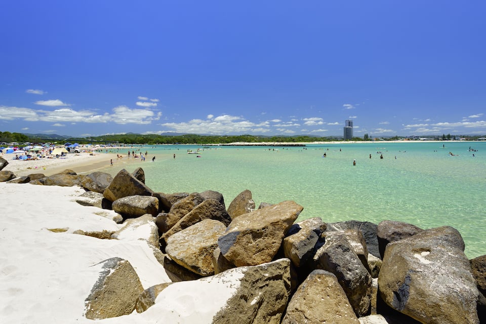 A beach with large rocks in the foreground, clear turquoise water, and a distant building.