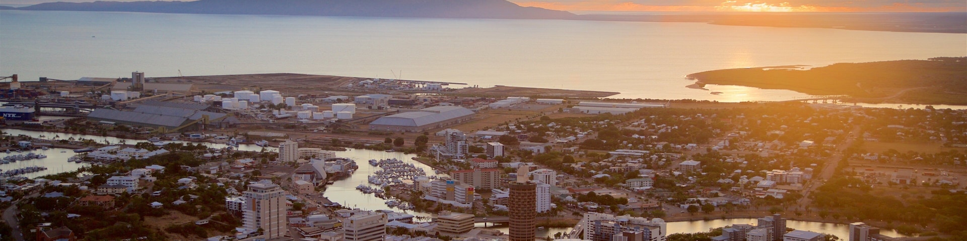 A coastal cityscape at sunset with a river, marina, and mountains in the background.