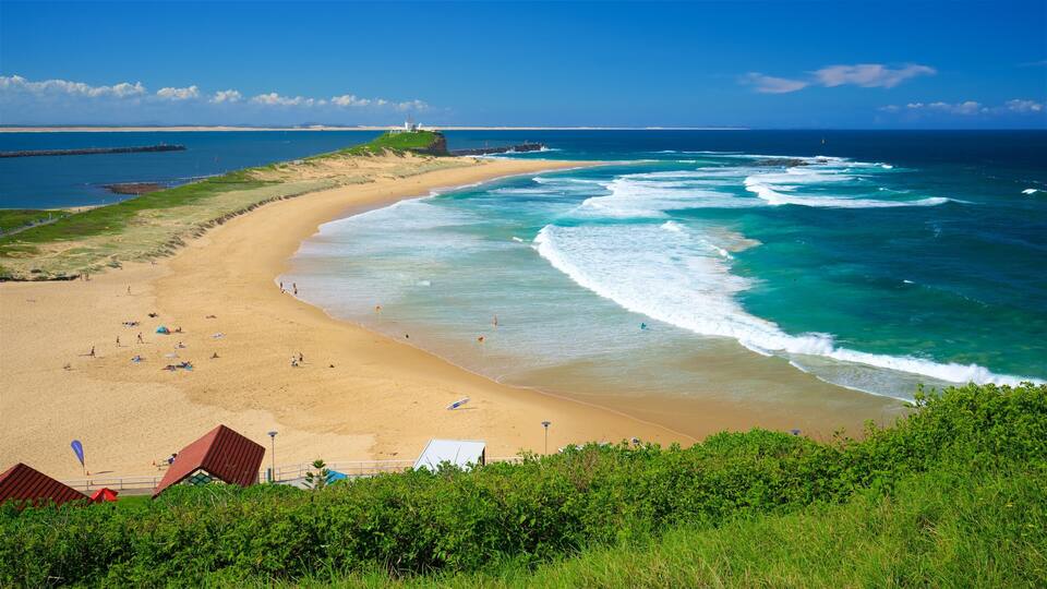 A sandy beach with waves, a lighthouse, and greenery.