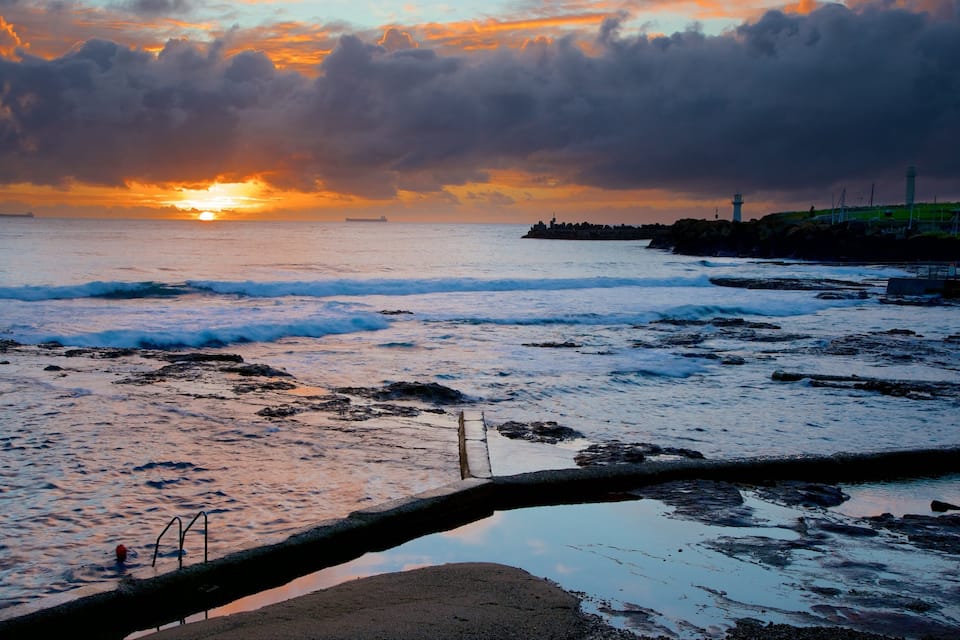 Un atardecer sobre una costa rocosa con olas, un faro y un barco distante.