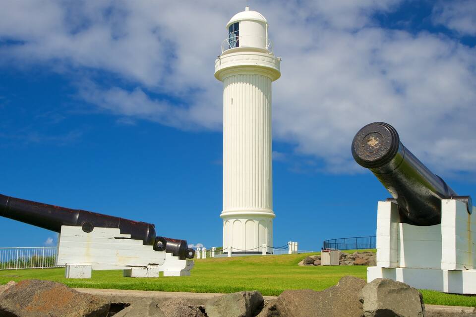 A lighthouse and two cannons on a grassy hill with a clear blue sky.