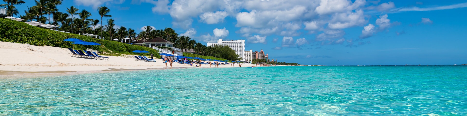 Una playa con aguas turquesas cristalinas, palmeras y un edificio blanco al fondo.