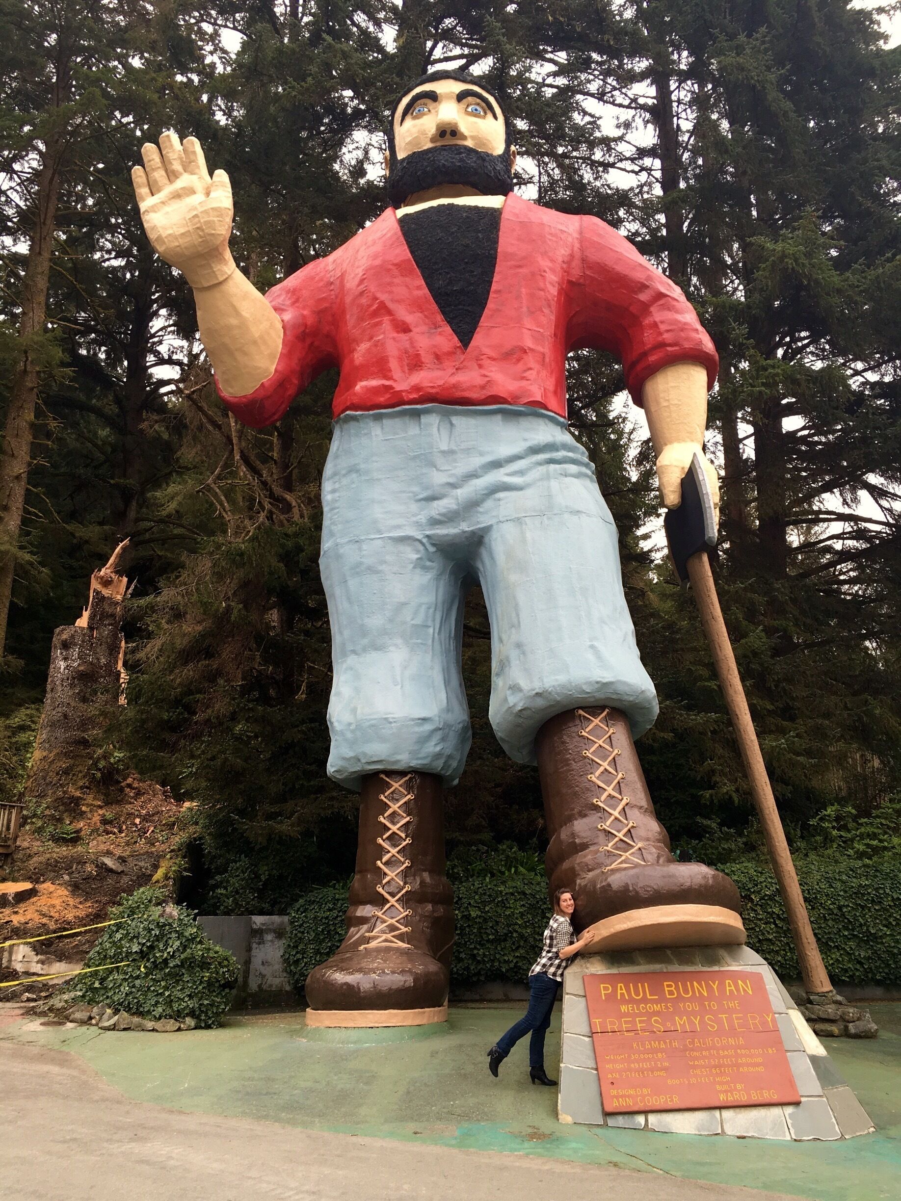 A large statue of a man with a raised hand, wearing a red shirt and blue pants, standing in a forested area with a plaque in front.