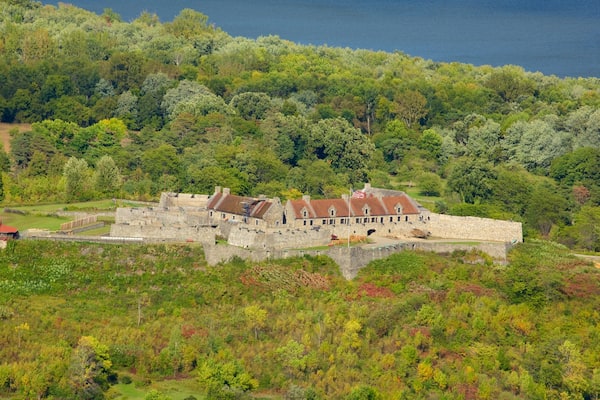 A historic stone building with a red roof, surrounded by lush greenery and a forested hill.