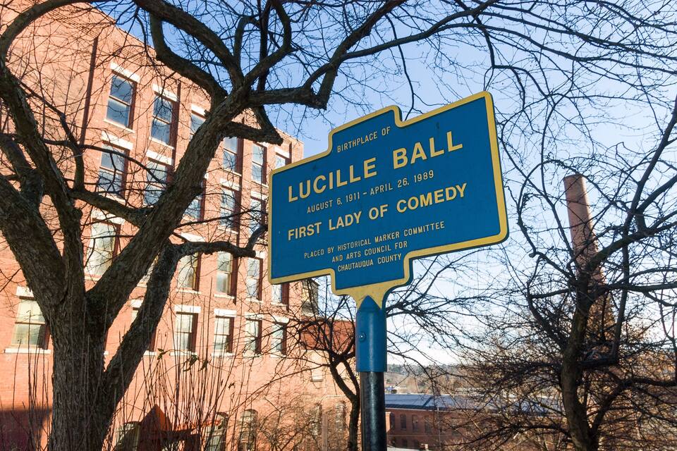 Placa azul de marco histórico com texto branco indicando "Lucille Ball, 6 de agosto de 1911 – 26 de abril de 1989, Primeira-Dama da Comédia, Instalada pelo Comitê de Marcos Históricos do Condado de Chautauqua."