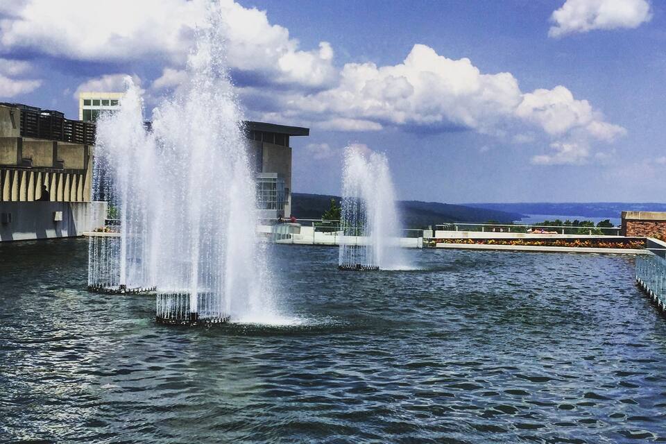A water fountain with multiple jets, a modern building, and a clear blue sky with clouds.