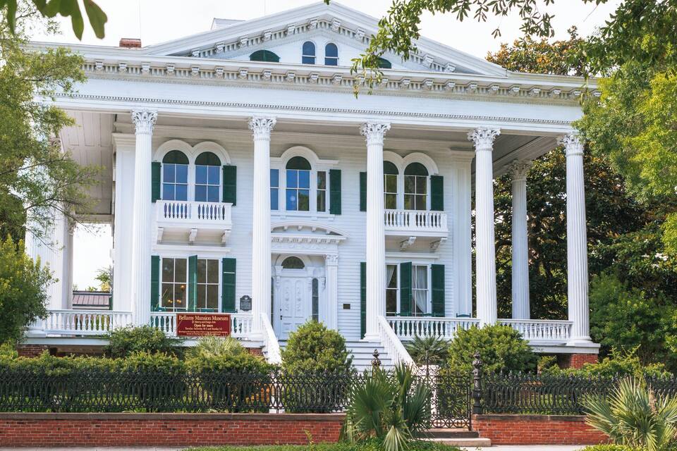 Um edifício de estilo colonial branco com persianas verdes, colunas e varanda.