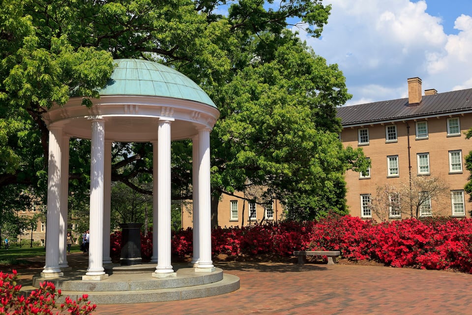 A gazebo with a green dome, surrounded by red flowers and trees, in front of a brick building.