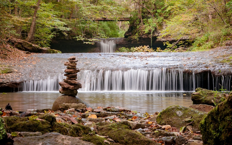 A serene forest with a small waterfall, a moss-covered rock stack, and a bridge in the background.