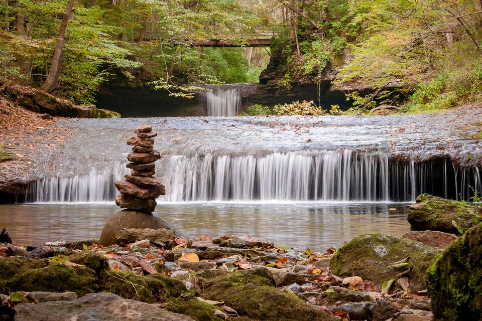 A serene forest with a small waterfall, a moss-covered rock stack, and a bridge in the background.