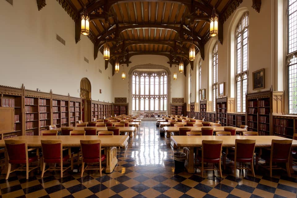 A large, well-lit library with high ceilings, wooden bookshelves, and checkered flooring.