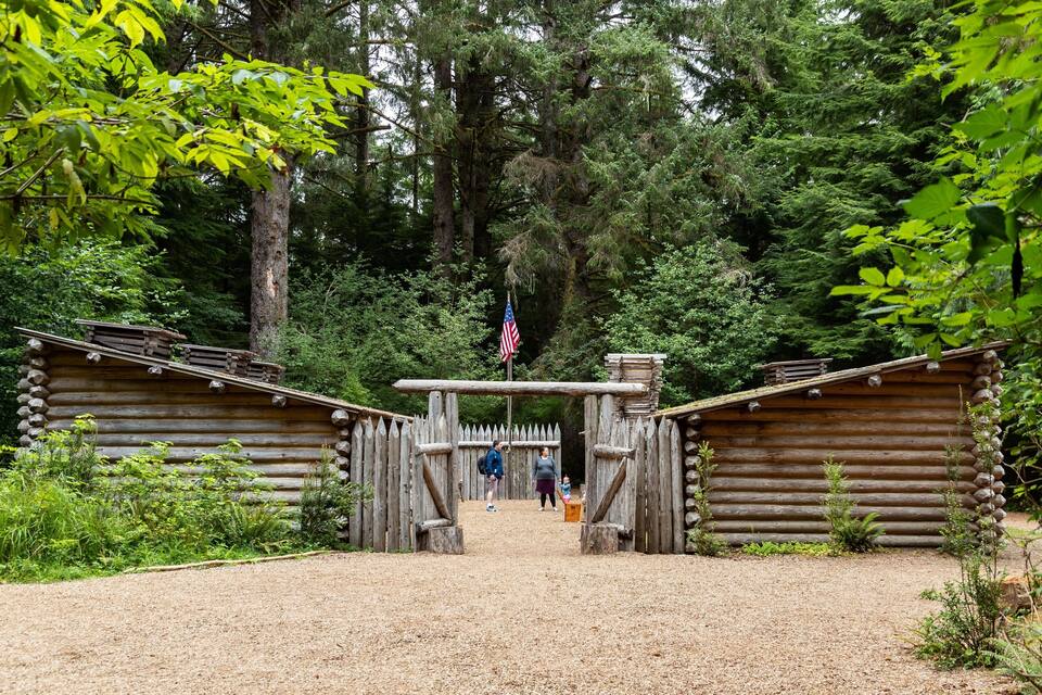 Une porte en bois rustique ornée d’un drapeau américain, entourée d’une forêt dense et de deux cabanes en rondins.