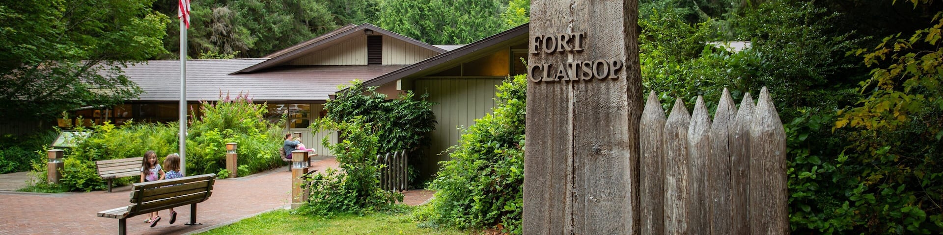 Un letrero de madera con la inscripción 'FORT CLATSO', rodeado de follaje y un edificio al fondo.