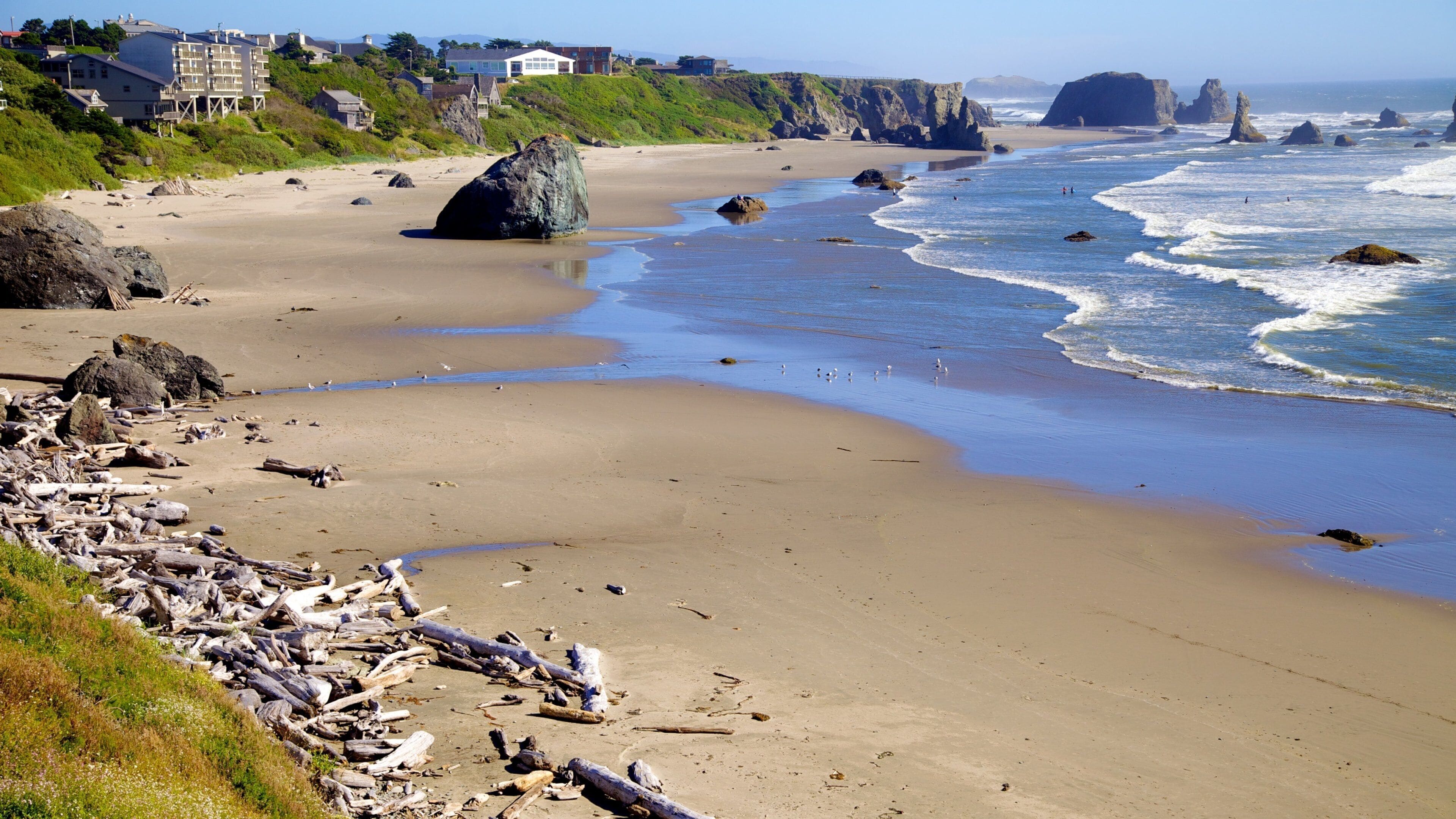A sandy beach with large rocks, waves, and a distant rocky coastline.