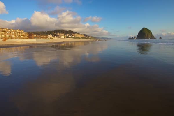A beach with a calm sea, a prominent green hill, and a row of buildings along the shore.