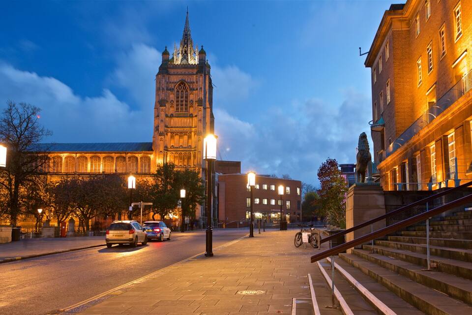 A city street at dusk with a prominent clock tower, a statue, and illuminated buildings.
