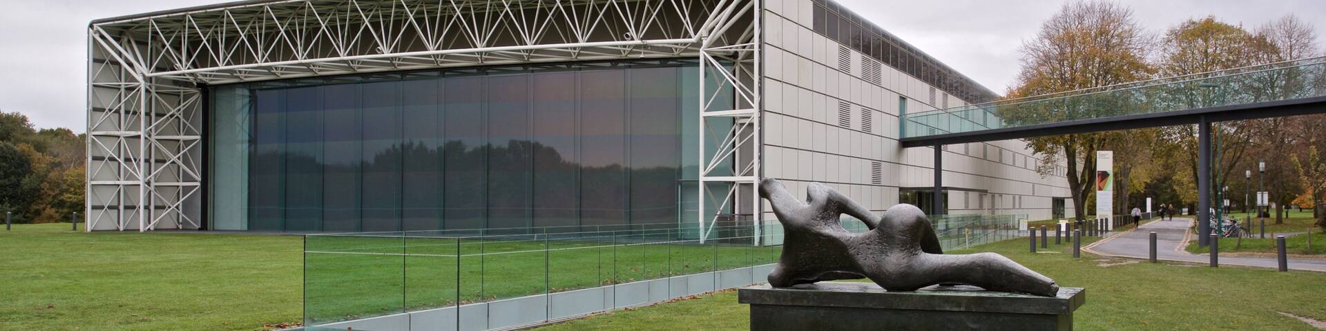 A modern building with a glass facade and a sculpture of a reclining figure in a park.