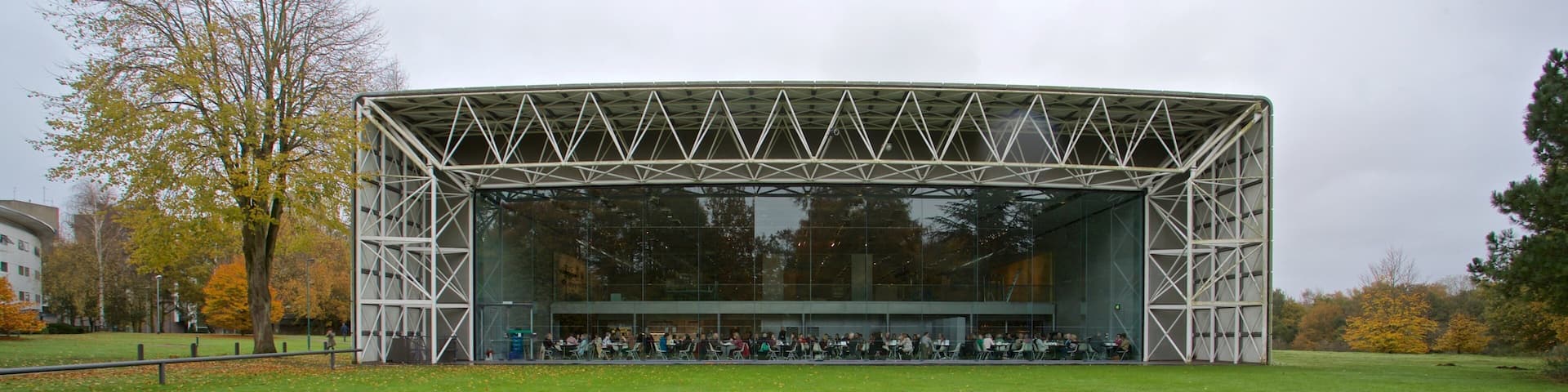 An open-air amphitheater with a metal frame and glass walls, surrounded by a grassy field and trees with autumn foliage.