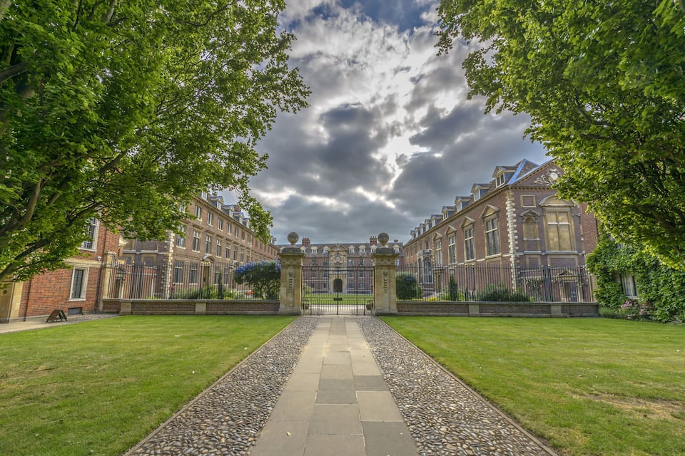 A historic building with a central courtyard, surrounded by lush green trees and a paved pathway leading to a gated entrance.